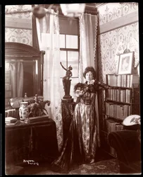 Anita Chartres steht in der Ecke eines Zimmers, ca. 1898